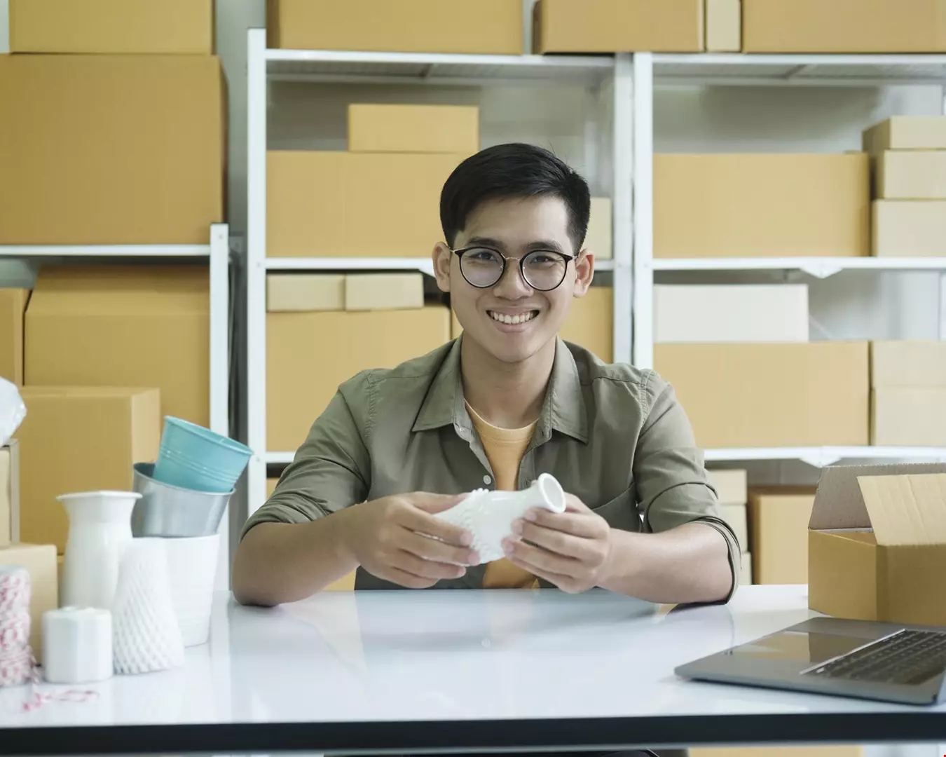 Ung man med glasögon sitter i lagermiljö och packar in beställningar i lådor har dator bredvid sig Akavia Aspekt